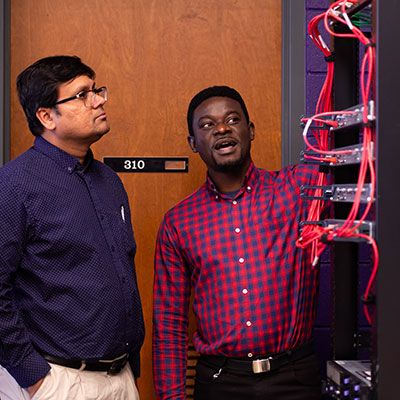 a student explaining something to a faculty member while standing in front of computer networking equipment