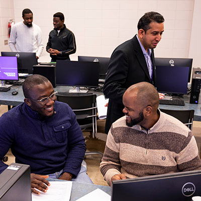 Two students smiling while talking about something at a computer. A professor and two other students are in the background.