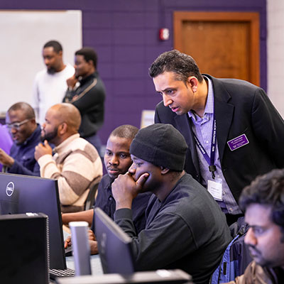 a row of students working in pairs at computers, while a professor helps one pair