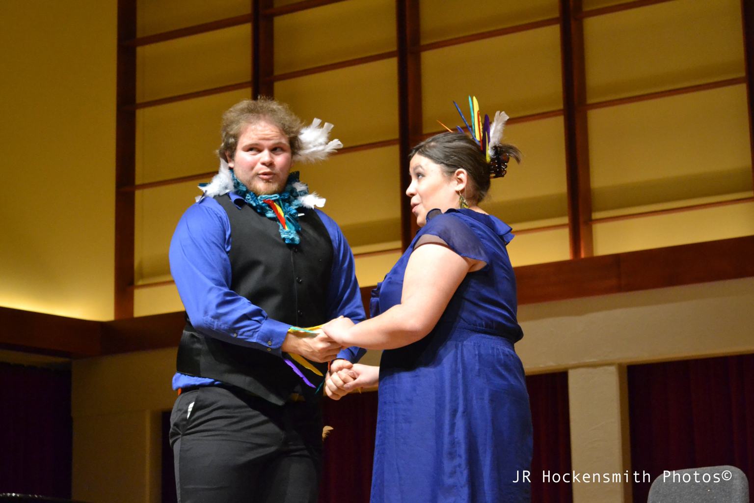 A man and woman adorned with feathers hold hands and sing a duet.