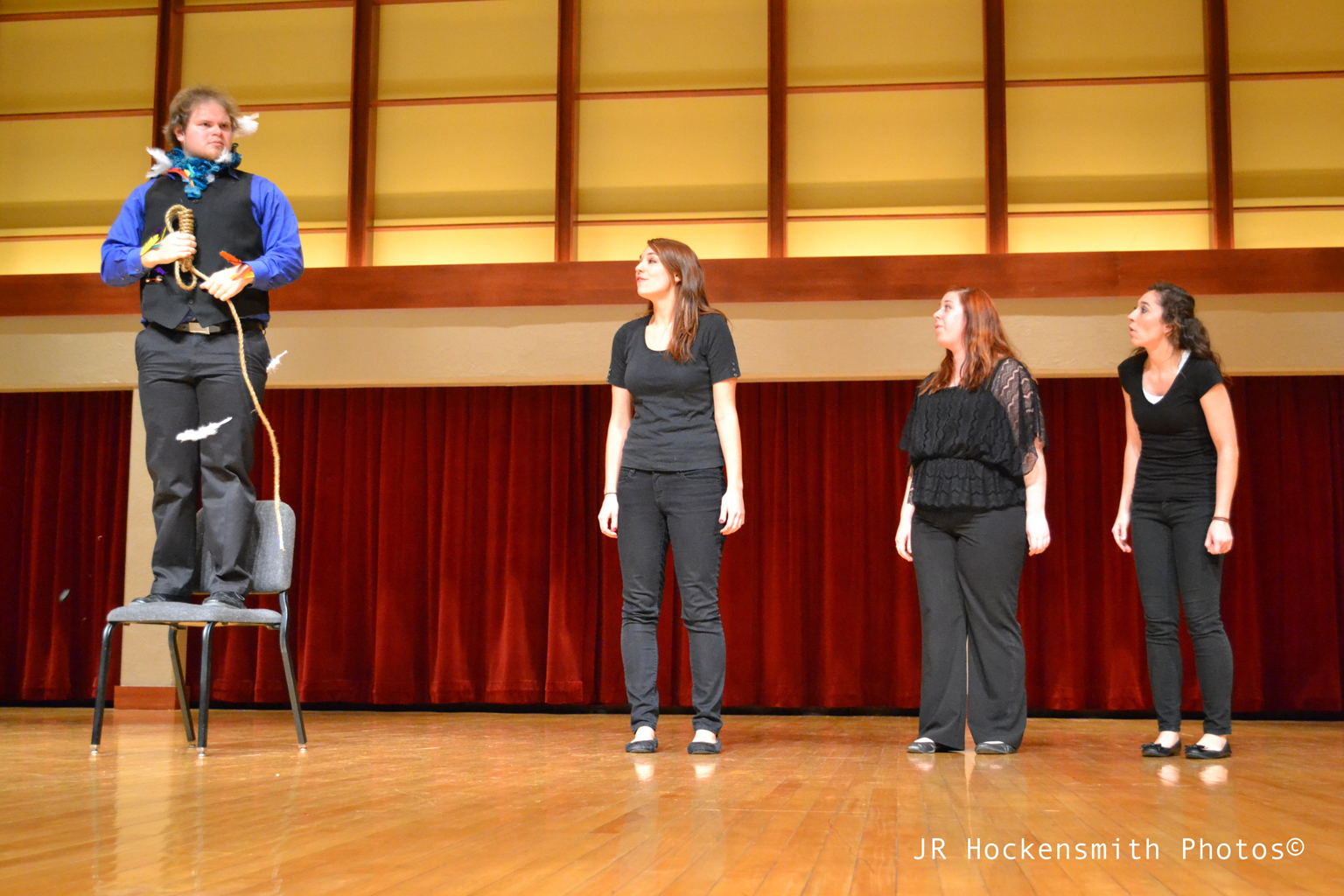 A man adorned with feathers stands on a chair and holds a noose while three women look on.