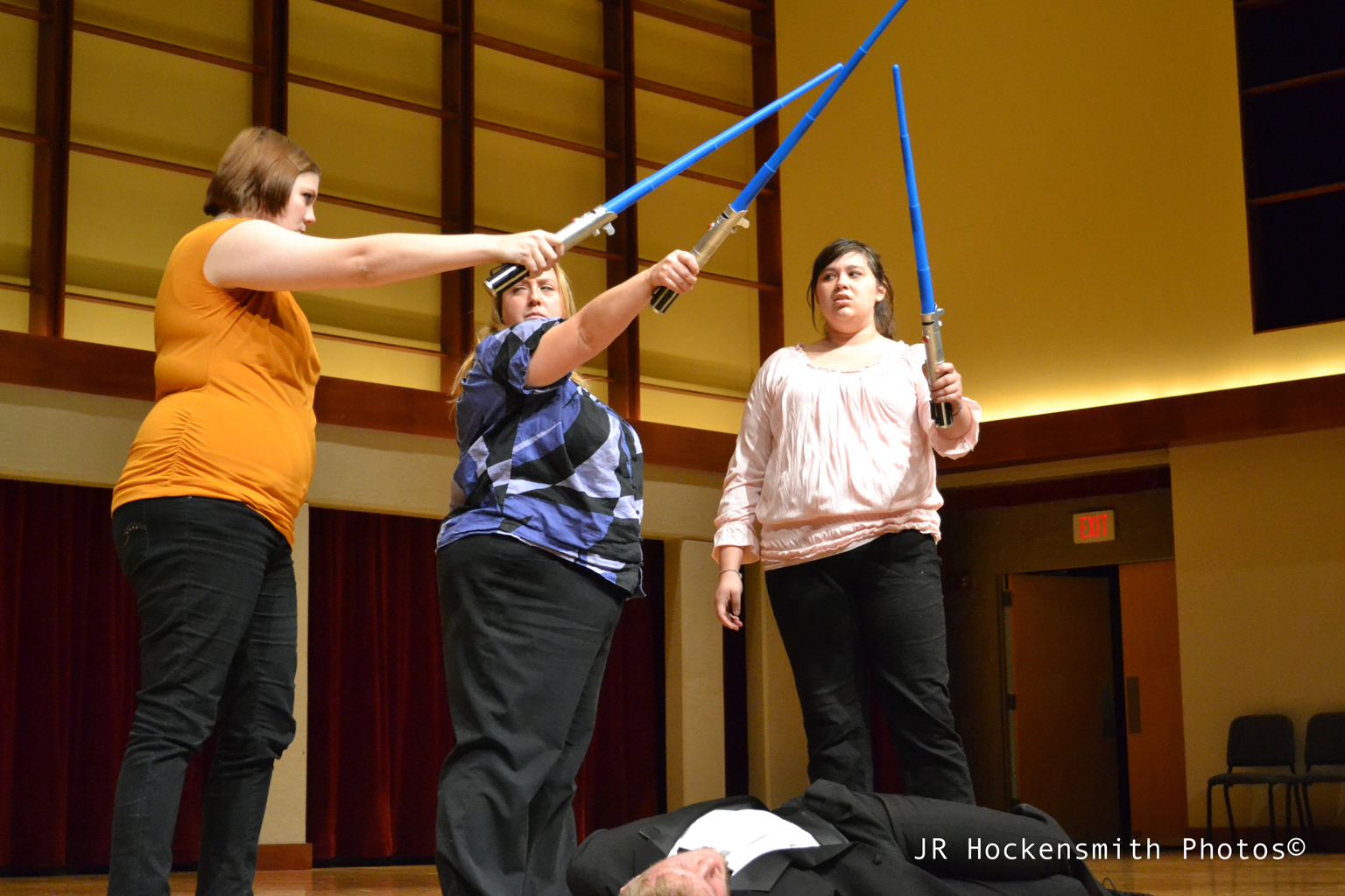 Three women holding light sabers stand over a man in a tuxedo lying on the ground.