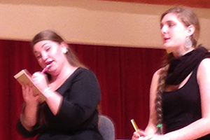 Two women sit in chairs. One woman holds a portrait of a man.